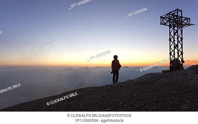 A hiker admires the sea of clouds from the summit cross of Punta Penia, Marmolada group, Dolomites, Canazei, Trento province, Trentino-Alto Adige, Italy