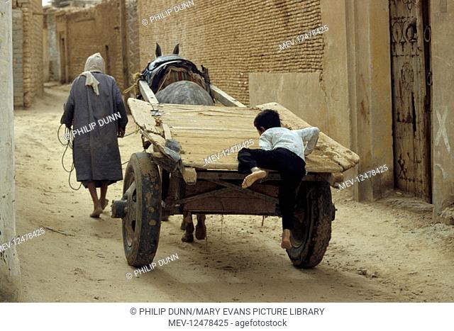 A boy jumps onto the back of a mule cart and gets a free ride down a dusty street. Nefta, Tunisia