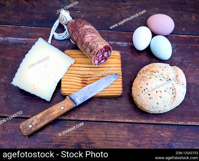 An Iberian pork sausage on an old wooden board with an antique knife, a loaf of Spanish rustic seed bread, cheese and three multicolor eggs