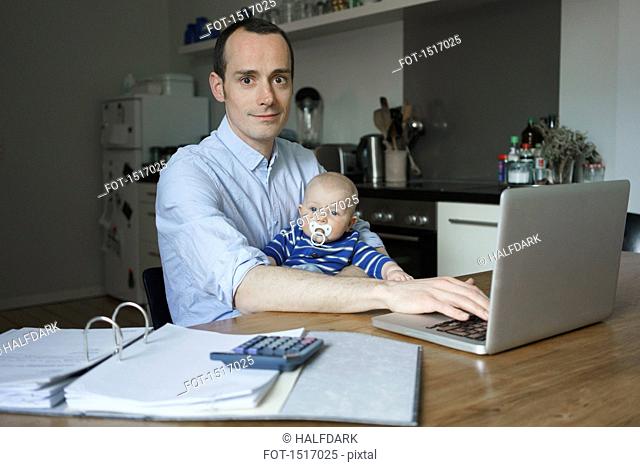 Portrait of father using laptop while carrying son at home