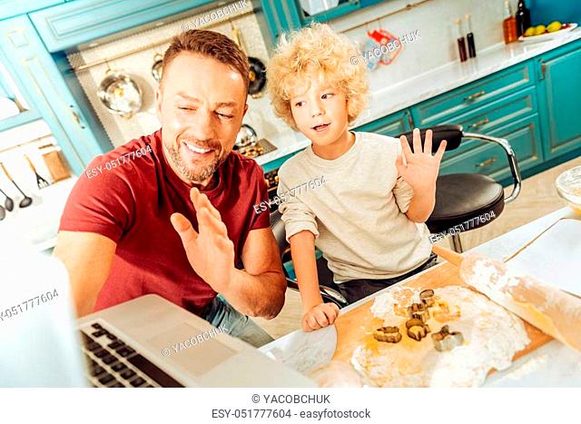 Say hello. Joyful nice positive man holding a laptop and waving his hand while standing in the kitchen together with his son