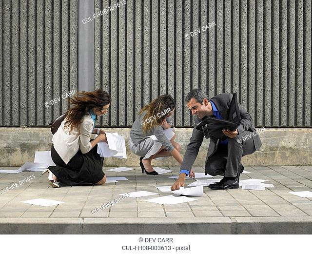 Two businesswomen and one businessman trying to recover papers blowing about pavement, Alicante, Spain