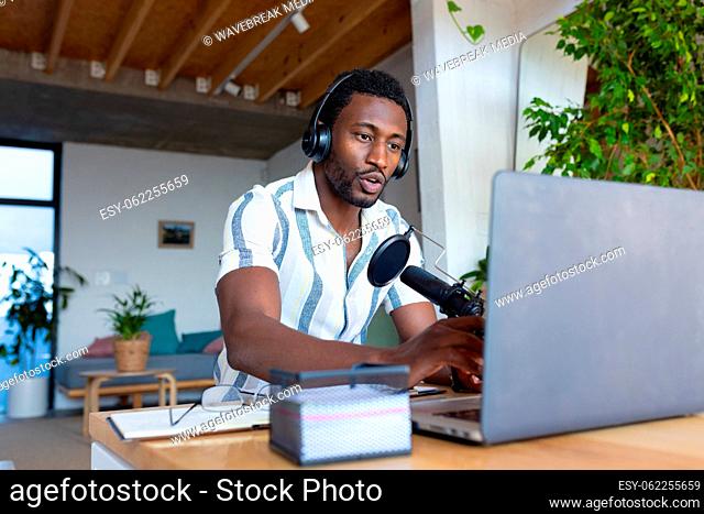 Happy african american man sitting at table in kitchen, using laptop and making vlog