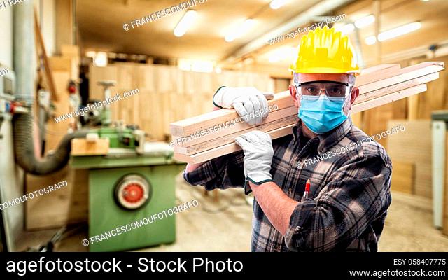 Carpenter worker at work in the carpentry workshop, wears helmet, goggles, leather gloves and surgical mask to prevent coronavirus infection