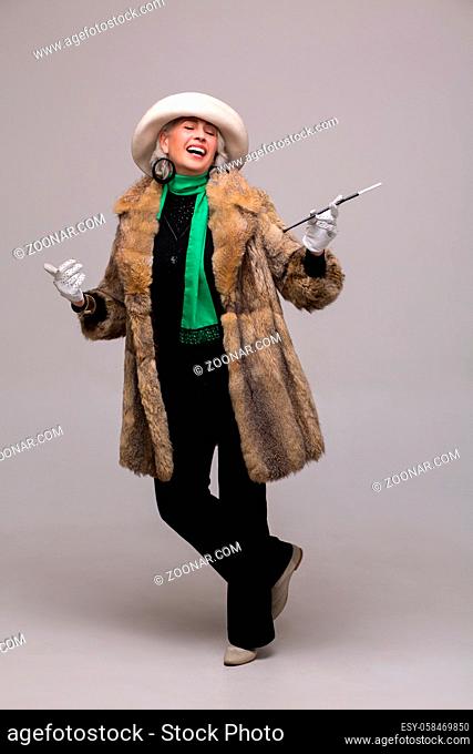 Senior rich woman smoking cigarette and smiling for camera. Fashionable lady in expensive fur posing in full length in studio
