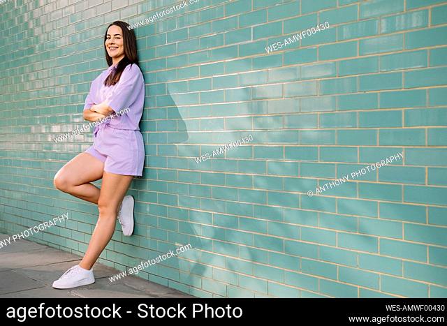 Happy young woman with arms crossed leaning on wall