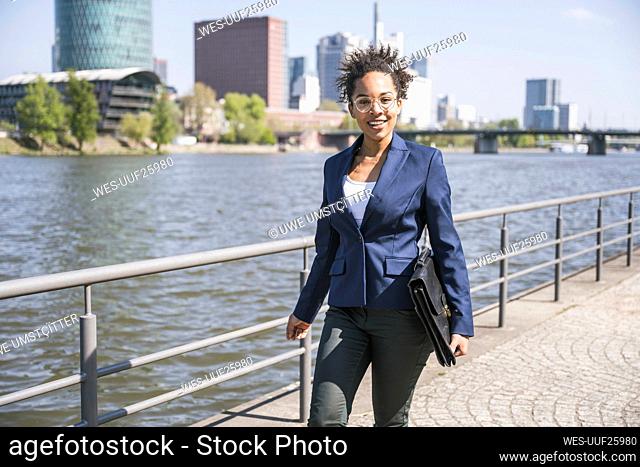Smiling businesswoman with briefcase walking by River Main on sunny day