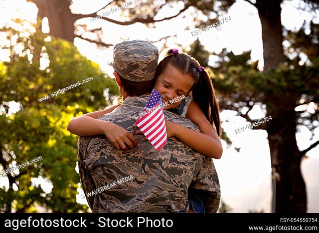 Rear view of a young adult mixed race male soldier in the garden outside his home, carrying his daughter, who is leaning over his shoulder holding a US flag and...