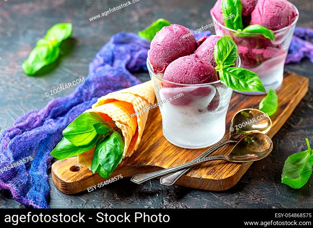 Artisanal blueberry ice cream in glasses with green basil on a wooden serving board on a dark textured background. Selective focus