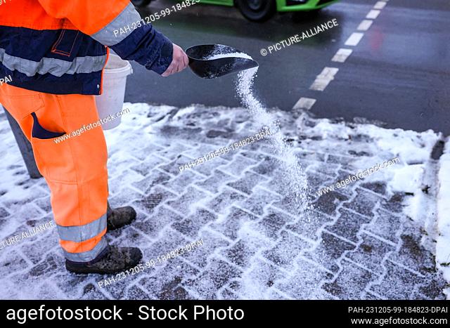 05 December 2023, Saxony, Leipzig: An employee of the winter road clearance service spreads road salt on the footpath at a road junction in the rain and...