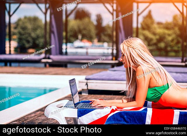 Blonde beautiful amazing woman working with laptop by the swimming pool. Working into pleasure. Blurred background