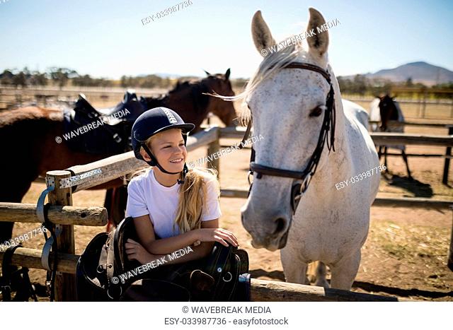 Smiling girl leaning on the fence and looking at the white horse in ranch