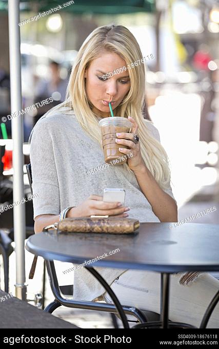 Young woman sitting outside at a cafe on her phone drinking iced coffee