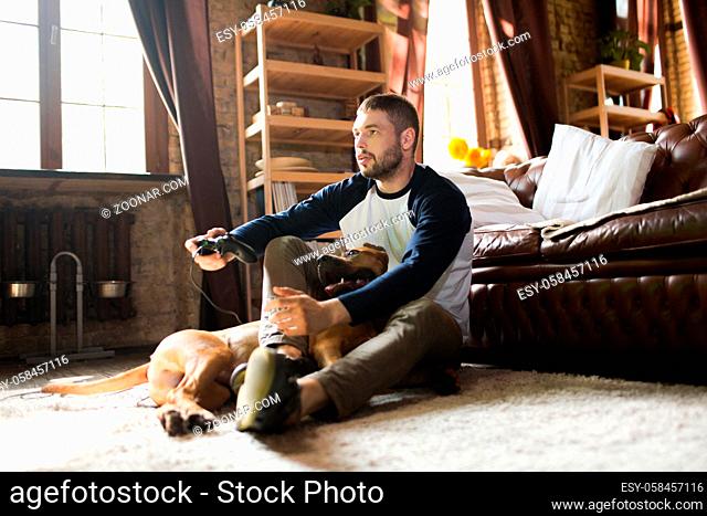 Man and dog on the floor at home. Male enjoying video game in a company with staffordshire terrier