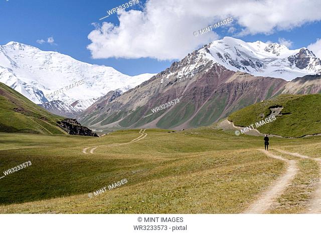 Man standing on tire tracks in a valley surrounded by snow capped mountains, Tulpar Kul, Kyrgyzstan, looking towards Peak Lenin