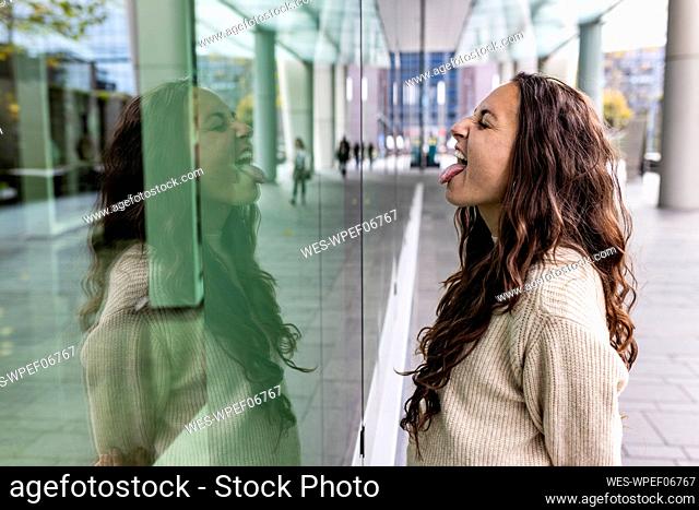 Happy woman sticking out tongue looking at reflection on glass wall