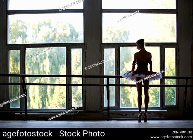 Elegant young ballerina standing near a large window in a dance class. Relying on the dance machine looks somewhere. Silhouette against a window