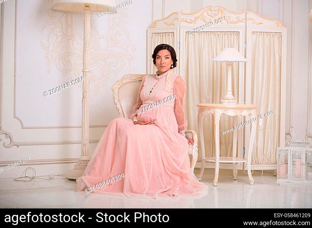 Portrait of brunette pregnant lady smiling and touching her belly while sitting in luxurious armchair in vintage interior in expensive apartments