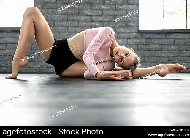 Cute ballerina in dance wear lies on the stretched left leg on a floor on the brick wall and windows background. Her right leg is on the toe