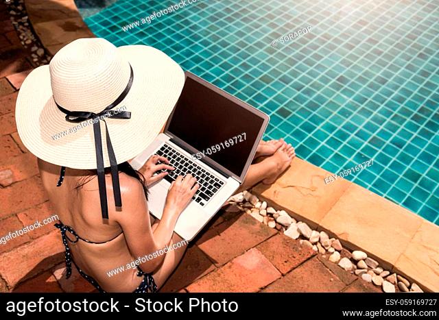Asian Young Woman using her laptop with big hat on poolside edge on sunny day blue water