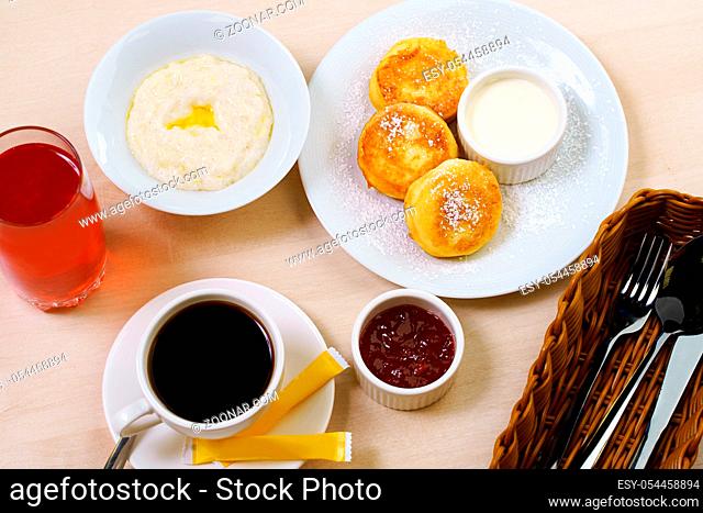 Table set for breakfast with rice porridge, cheesecakes with jam, berry drink, black coffee and cuttlery in wooden container in cafe