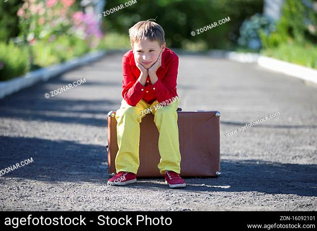 A sad boy sits on an old suitcase on the road. The child is about to leave