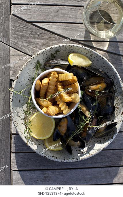Mussels in a white wine, lemon, garlic and thyme sauce, with french fries, lemon slices, and a glass of white wine, on an outdoor table