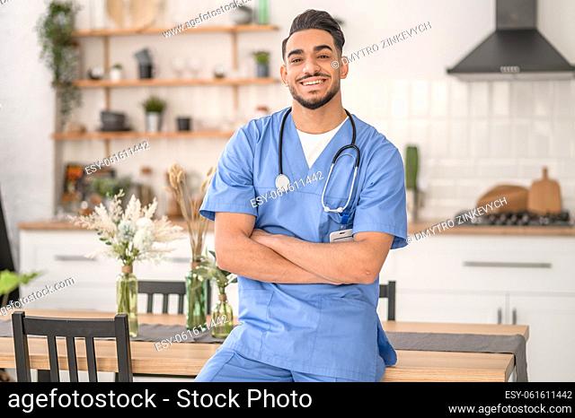 Smiling joyous young healthcare worker seated on the edge of the table looking before him