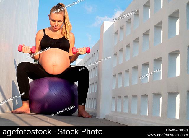 Portrait of pretty pregnant woman exercises with fitball and dumbbells outdoors. Working out and fitness, pregnancy concept