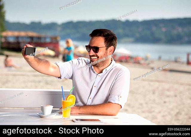 Handsome man with sunglasses using phone for web chatting on beach. Enjoying beside river