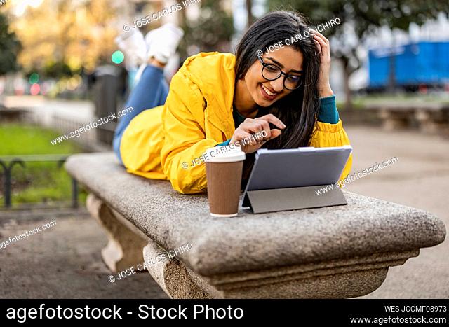 Happy young woman using tablet PC lying on bench