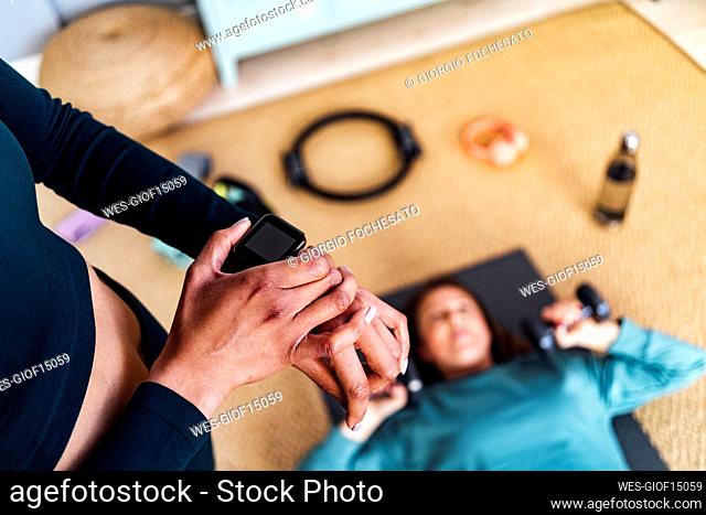 Young woman adjusting smart watch by friend exercising at home