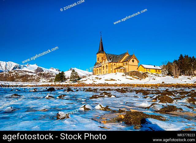The wooden yellow Vågan Church on the beach on the Lofoten islands in Norway in winter with frozen river and mountains in morning light