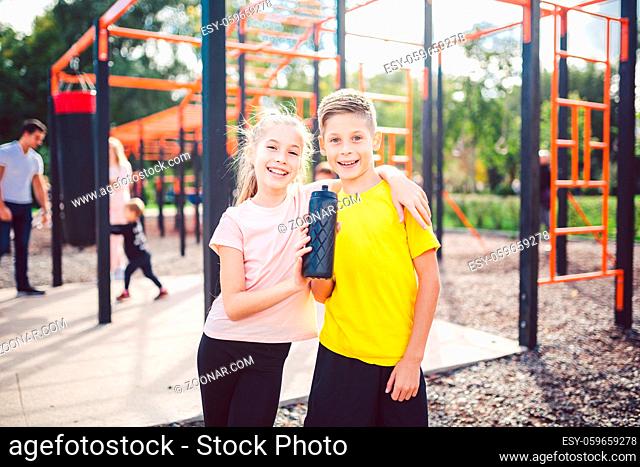 Caucasian children brother and sister stand in embrace at outdoor sports ground with smile and flask with water after training