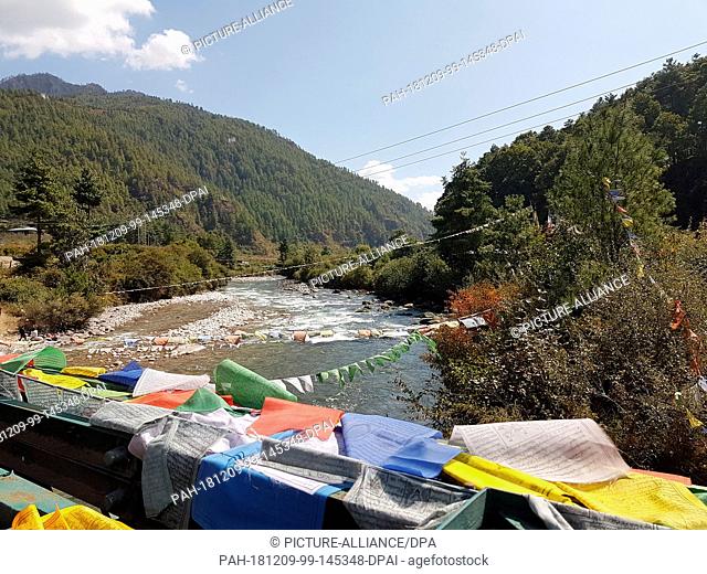 17 October 2018, Bhutan, Paro: Prayer flags and power lines cross a river in the Paro Valley. Bhutan is considered the only climate-neutral country in the world