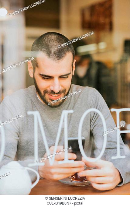 Young man sitting in cafe, using smartphone, view through window
