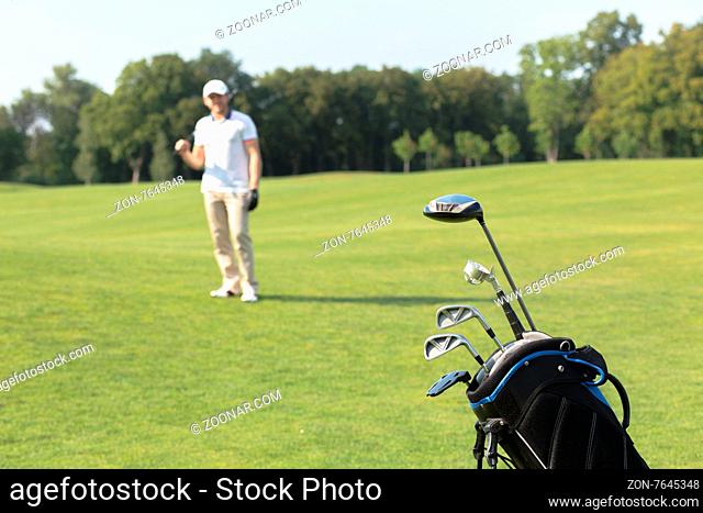 Golf game concept. Golf clubs in bag against the golf course. Golfer standing on the background and holding a driver in his hand