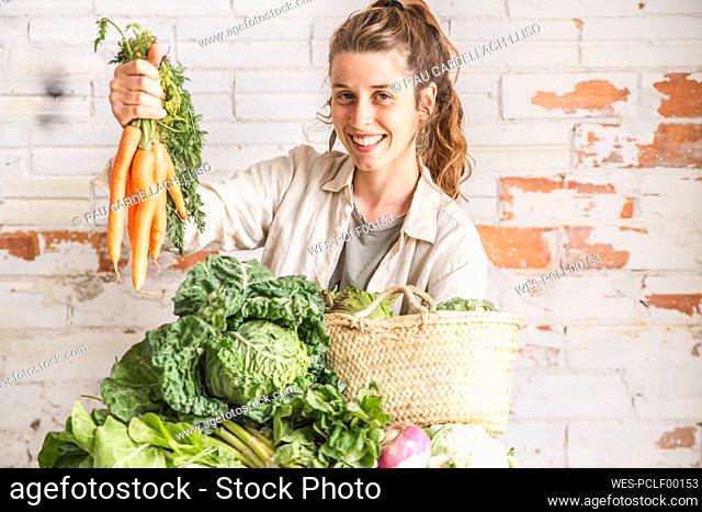 Happy grocer holding bunch of carrots in front of brick wall