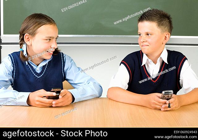 Smiling, young boy and girl sitting at desk and holding mobile phones. Looking at each other. Front view