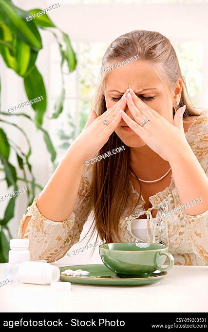 Young woman having headache, holding her head, leaning on table. Taking pills with tea