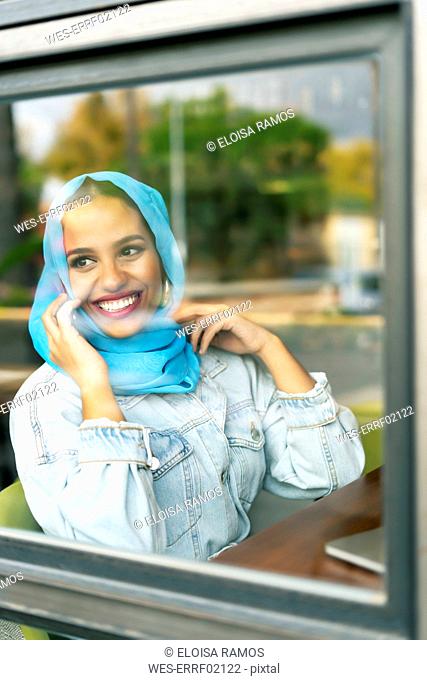 Young woman wearing turquoise hijab and phoning in a cafe, looking through window