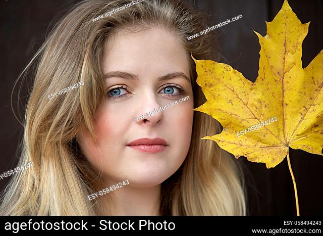 Close up is the face of a beautiful girl holding a yellow maple leaf. Blue-eyed blonde in autumn