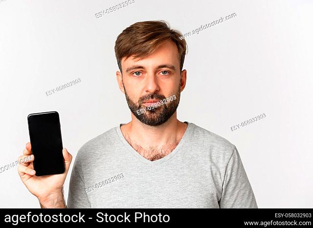 Close-up of tired caucasian guy in gray t-shirt, looking exhausted and showing smartphone screen, standing over white background