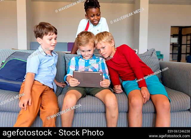 Multiracial elementary schoolboys looking at digital tablet while sitting on couch in school