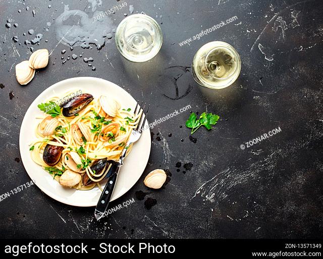 Spaghetti vongole, Italian seafood pasta with clams and mussels, in plate with herbs and two glasses of white wine on rustic stone background, space for text