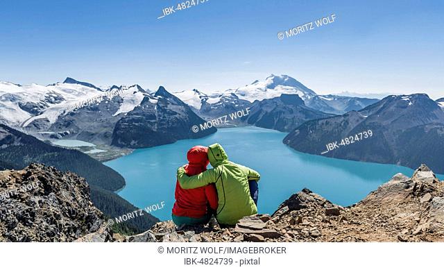 View from Panorama Ridge trail, Two hikers sitting on a rock with Garibaldi Lake, turquoise glacial lake, Guard Mountain and Deception Peak, back glacier