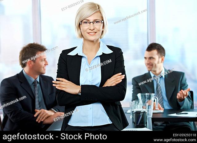 Portrait of smiling businesswoman standing with folded arms at meeting with sitting coworkers
