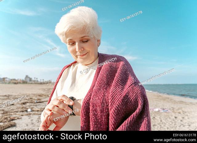Waist-up portrait of a smiling pleased senior woman looking at the fitness tracker on her wrist