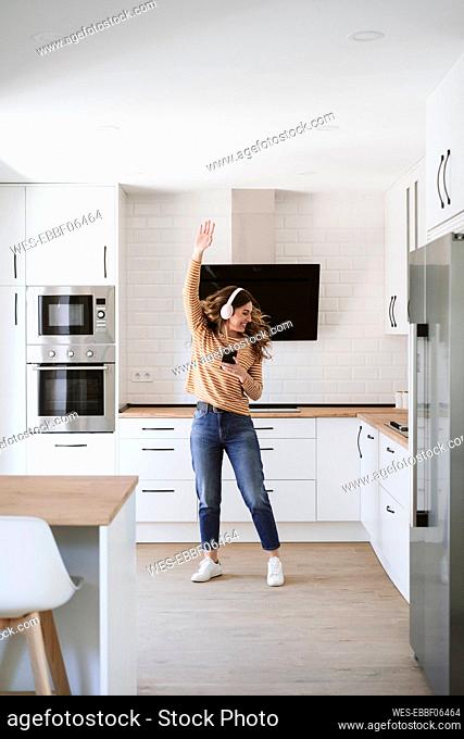 Cheerful young woman listening to music with headphones in kitchen at home