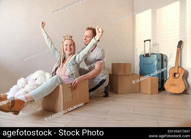 Happy young couple moving to a new apartment. A man pushes a woman sitting in a box with arms raised. Packed things in the background